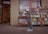 A bookstore with wooden shelves filled with various books placed outside the store. There's a neon sign that reads 'BOOKS' in the window. In the foreground, there is a red metal cart with books. The store door is open, revealing more shelves and books inside, with an 'OPEN' sign visible. A blurred figure is seen walking past the store, indicating some motion.