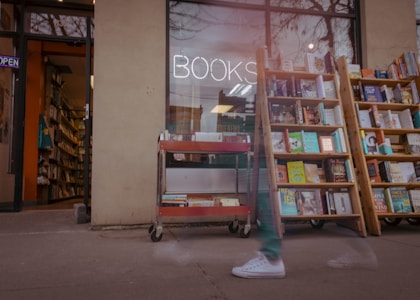 A bookstore with wooden shelves filled with various books placed outside the store. There's a neon sign that reads 'BOOKS' in the window. In the foreground, there is a red metal cart with books. The store door is open, revealing more shelves and books inside, with an 'OPEN' sign visible. A blurred figure is seen walking past the store, indicating some motion.