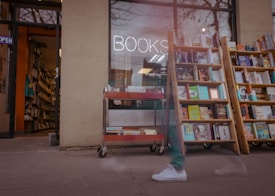 A bookstore with wooden shelves filled with various books placed outside the store. There's a neon sign that reads 'BOOKS' in the window. In the foreground, there is a red metal cart with books. The store door is open, revealing more shelves and books inside, with an 'OPEN' sign visible. A blurred figure is seen walking past the store, indicating some motion.