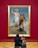 A large framed painting on a red wall depicts a biblical scene with two figures. In the foreground, two people are seated on the floor observing the artwork, with one wearing a backpack.
