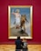 A large framed painting on a red wall depicts a biblical scene with two figures. In the foreground, two people are seated on the floor observing the artwork, with one wearing a backpack.