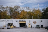 A white building labeled 'Brewery' is set against a backdrop of autumn trees. The right side of the building features a logo with a yellow door beneath it. In front of the entrance, there are small potted plants. To the left, an open garage door reveals storage items and equipment, including a forklift and several pallets. The surrounding area has gravel and some scattered objects, including a bicycle leaned against the building and a parked car partially visible on the right.