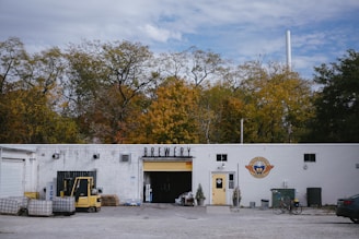 A white building labeled 'Brewery' is set against a backdrop of autumn trees. The right side of the building features a logo with a yellow door beneath it. In front of the entrance, there are small potted plants. To the left, an open garage door reveals storage items and equipment, including a forklift and several pallets. The surrounding area has gravel and some scattered objects, including a bicycle leaned against the building and a parked car partially visible on the right.