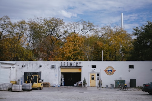 A white building labeled 'Brewery' is set against a backdrop of autumn trees. The right side of the building features a logo with a yellow door beneath it. In front of the entrance, there are small potted plants. To the left, an open garage door reveals storage items and equipment, including a forklift and several pallets. The surrounding area has gravel and some scattered objects, including a bicycle leaned against the building and a parked car partially visible on the right.