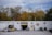 A white building labeled 'Brewery' is set against a backdrop of autumn trees. The right side of the building features a logo with a yellow door beneath it. In front of the entrance, there are small potted plants. To the left, an open garage door reveals storage items and equipment, including a forklift and several pallets. The surrounding area has gravel and some scattered objects, including a bicycle leaned against the building and a parked car partially visible on the right.