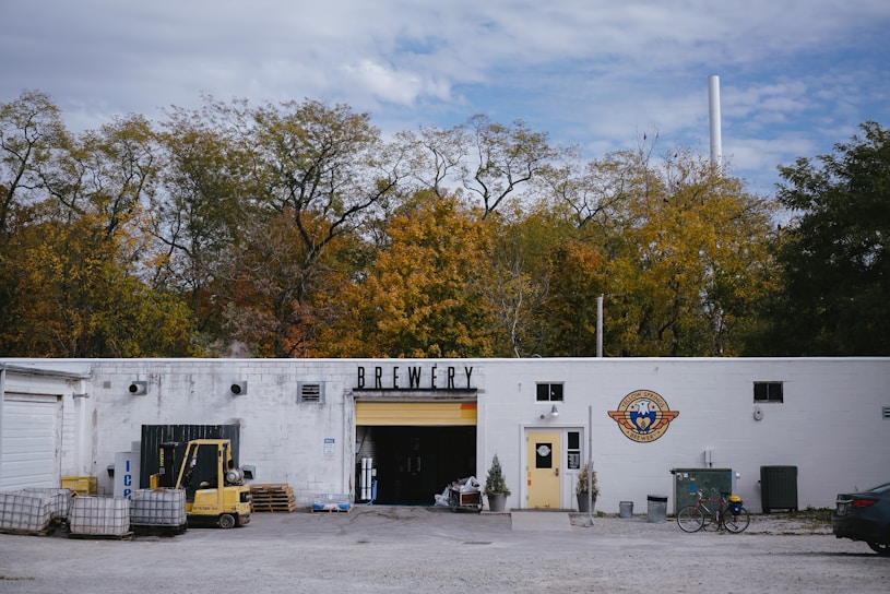 A white building labeled 'Brewery' is set against a backdrop of autumn trees. The right side of the building features a logo with a yellow door beneath it. In front of the entrance, there are small potted plants. To the left, an open garage door reveals storage items and equipment, including a forklift and several pallets. The surrounding area has gravel and some scattered objects, including a bicycle leaned against the building and a parked car partially visible on the right.