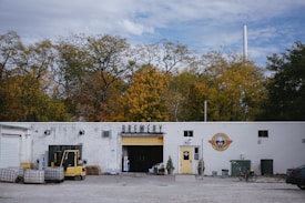 A white building labeled 'Brewery' is set against a backdrop of autumn trees. The right side of the building features a logo with a yellow door beneath it. In front of the entrance, there are small potted plants. To the left, an open garage door reveals storage items and equipment, including a forklift and several pallets. The surrounding area has gravel and some scattered objects, including a bicycle leaned against the building and a parked car partially visible on the right.