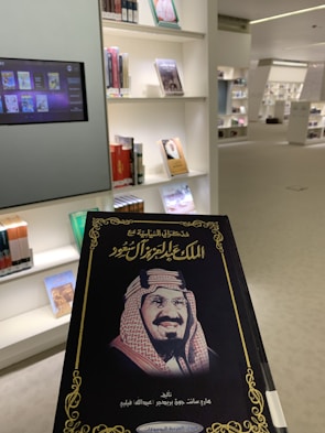 A book is being held up in a library. The cover features an image of a man in traditional attire and text in Arabic. Shelves filled with various books and a digital screen displaying book covers appear in the background.