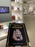 A book is being held up in a library. The cover features an image of a man in traditional attire and text in Arabic. Shelves filled with various books and a digital screen displaying book covers appear in the background.
