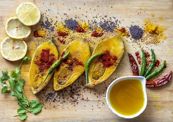 Three slices of raw fish marinated with turmeric and red chili powder are placed on a wooden cutting board. Around them are various spices, including mustard seeds and turmeric powder, and a few green and dried red chilies. Two slices of lemon and a bunch of cilantro are on the side, and a bowl containing yellow oil completes the arrangement.