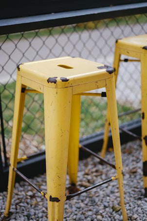 A yellow metal bar stool with visible wear and rust spots on the edges is positioned on gravel near a chain-link fence. Another similar stool is partially visible in the background. The surrounding area is outdoors with some greenery beyond the fence.