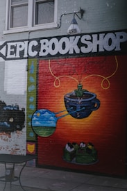 A mural on a brick wall features the words 'EPIC BOOKSHOP' in large white letters. Below, there is a colorful painting of a cup with green tendrils emerging, leading to artistic depictions of planets and a landscape within bubbles. The background transitions from dark red to orange, creating a vibrant contrast. Above the mural, a lamp is mounted on the wall.