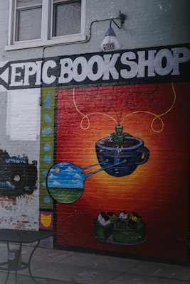 A mural on a brick wall features the words 'EPIC BOOKSHOP' in large white letters. Below, there is a colorful painting of a cup with green tendrils emerging, leading to artistic depictions of planets and a landscape within bubbles. The background transitions from dark red to orange, creating a vibrant contrast. Above the mural, a lamp is mounted on the wall.