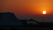 The image captures a stunning sunset over an industrial landscape, with the sun setting against a vibrant orange sky. A large mound of material, possibly salt or sand, is visible on the left. In the foreground, there are silhouettes of industrial structures, including conveyor belts and metal frameworks.