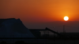 The image captures a stunning sunset over an industrial landscape, with the sun setting against a vibrant orange sky. A large mound of material, possibly salt or sand, is visible on the left. In the foreground, there are silhouettes of industrial structures, including conveyor belts and metal frameworks.