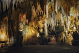 Intricate limestone formations hang from the ceiling of a dimly lit cave, creating a breathtaking display of stalactites and stalagmites. Warm yellow lighting highlights the natural textures and contours of the rock formations, casting dramatic shadows across the scene. In the center, a small wooden organ is positioned, adding a unique and unexpected element to the subterranean environment.