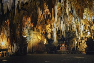 Intricate limestone formations hang from the ceiling of a dimly lit cave, creating a breathtaking display of stalactites and stalagmites. Warm yellow lighting highlights the natural textures and contours of the rock formations, casting dramatic shadows across the scene. In the center, a small wooden organ is positioned, adding a unique and unexpected element to the subterranean environment.