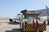 A large airplane is parked on the tarmac near a boarding gate. In front of the aircraft, a tow vehicle is visible, painted with black and yellow diagonal stripes, indicating it's in use for ground support. The terminal gate number G1 is clearly marked, and the background includes a clear blue sky.