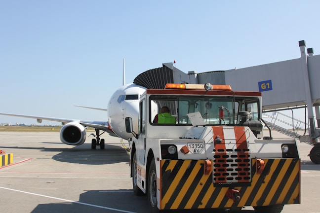 A large airplane is parked on the tarmac near a boarding gate. In front of the aircraft, a tow vehicle is visible, painted with black and yellow diagonal stripes, indicating it's in use for ground support. The terminal gate number G1 is clearly marked, and the background includes a clear blue sky.