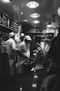 A black and white image of an interior restaurant scene featuring staff wearing uniforms and hats, busy with food preparation behind a counter. The ceiling has round lights, and the wall displays menus. The atmosphere reflects an old-fashioned diner or fish and chips shop.