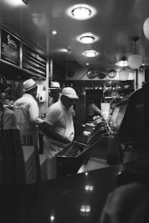 A black and white image of an interior restaurant scene featuring staff wearing uniforms and hats, busy with food preparation behind a counter. The ceiling has round lights, and the wall displays menus. The atmosphere reflects an old-fashioned diner or fish and chips shop.