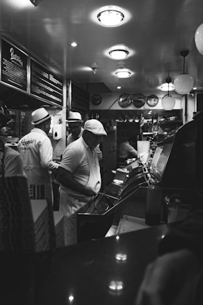 A black and white image of an interior restaurant scene featuring staff wearing uniforms and hats, busy with food preparation behind a counter. The ceiling has round lights, and the wall displays menus. The atmosphere reflects an old-fashioned diner or fish and chips shop.