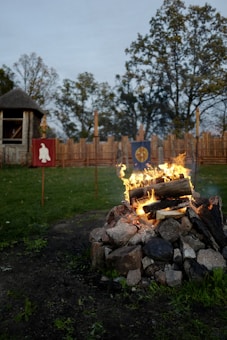 A bonfire surrounded by stones with burning logs is set in an open grassy area. In the background, there are wooden fences with medieval-style banners hanging, one red with a white symbol and another blue with a yellow design. Trees with sparse leaves line the area, and a small thatched-roof structure is visible to the left.
