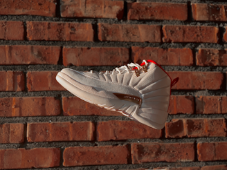 A white sneaker with red accents is prominently displayed against a rustic red brick wall, giving a sense of motion as it appears suspended in mid-air. The shoe features detailed stitching and gold lace eyelets, highlighting its design.