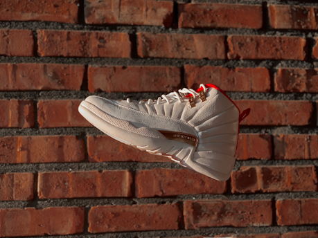 A white sneaker with red accents is prominently displayed against a rustic red brick wall, giving a sense of motion as it appears suspended in mid-air. The shoe features detailed stitching and gold lace eyelets, highlighting its design.