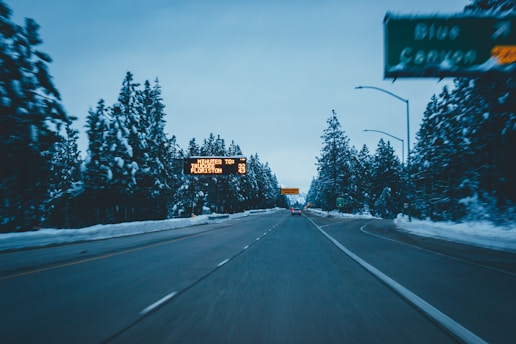A wintery scene on a highway with snow-covered pine trees on both sides. An electronic road sign displays travel information to nearby towns, and a freeway exit sign is visible. The sky is overcast, contributing to a cold, subdued atmosphere.