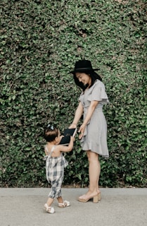 A woman in a striped dress and black hat is interacting with a young child in a plaid outfit, standing in front of a lush green wall of leaves. The woman is bending slightly and smiling, while the child reaches up holding a book.