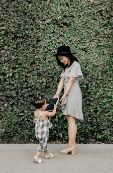 A woman in a striped dress and black hat is interacting with a young child in a plaid outfit, standing in front of a lush green wall of leaves. The woman is bending slightly and smiling, while the child reaches up holding a book.