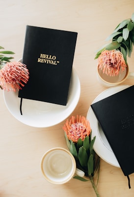 A flat lay composition featuring two black notebooks with gold embossed lettering titled 'HELLO REVIVAL' on a light wooden surface. Accompanying the notebooks are two vibrant orange and pink protea flowers with green leaves, placed in simple ceramic vases. A white ceramic cup and plates are also part of the arrangement, creating a minimalist and aesthetically pleasing setup.