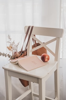 A white wooden chair is placed in front of a softly lit background. On the seat of the chair, there is an open book with a beige cover, a peach, dried flowers, and a brown bag hanging from the backrest. The overall arrangement is minimalistic and aesthetically pleasing.