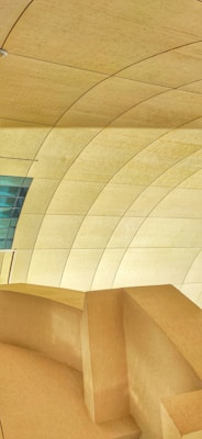 A modern architectural interior featuring a series of large, curved, beige panelled walls and ceiling. The design includes sharp lines and smooth curves, creating an organic and futuristic appearance. A small section of blue-tinted glass panels adds contrast to the predominantly warm color scheme.