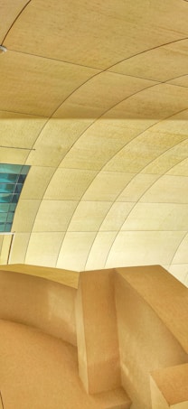 A modern architectural interior featuring a series of large, curved, beige panelled walls and ceiling. The design includes sharp lines and smooth curves, creating an organic and futuristic appearance. A small section of blue-tinted glass panels adds contrast to the predominantly warm color scheme.