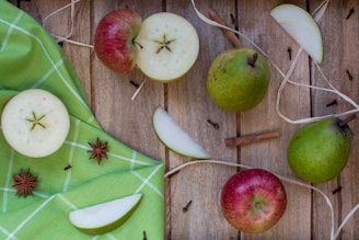 A rustic setup featuring whole and sliced apples and pears on a wooden surface. Cinnamon sticks, star anise, and a few cloves add a fragrant touch, while a green checkered cloth adds a pop of color to the arrangement.