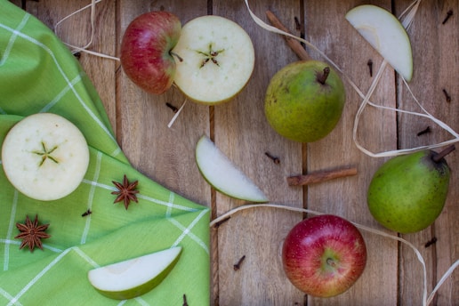 A rustic setup featuring whole and sliced apples and pears on a wooden surface. Cinnamon sticks, star anise, and a few cloves add a fragrant touch, while a green checkered cloth adds a pop of color to the arrangement.