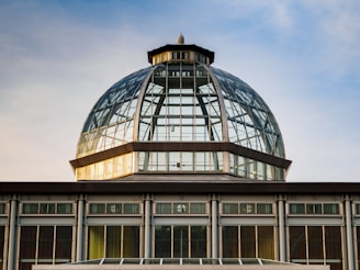 A large glass dome structure with a central spire, composed of metal frameworks and large panes of glass. The dome sits atop a rectangular building with evenly spaced vertical windows, reflecting golden sunlight under a blue sky.