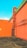 A vibrant outdoor scene featuring a bold orange wall with an industrial-style metal pipe attached to it. The wall extends to form a corner, and a metal gate provides an entrance at the base of the corner. The roof has a green section, contrasting with the orange walls. The ground is paved, and there are parking indicators with the number 'P5' visible on the wall.