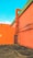 A vibrant outdoor scene featuring a bold orange wall with an industrial-style metal pipe attached to it. The wall extends to form a corner, and a metal gate provides an entrance at the base of the corner. The roof has a green section, contrasting with the orange walls. The ground is paved, and there are parking indicators with the number 'P5' visible on the wall.