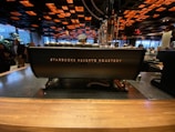 A Starbucks Reserve Roastery counter, featuring a large coffee machine with the brand's logo. The interior has a modern, industrial design with a dark ceiling adorned with orange and black square patterns. Several people are visible in the background, creating a lively cafe atmosphere.