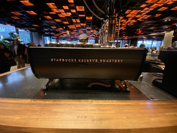 A Starbucks Reserve Roastery counter, featuring a large coffee machine with the brand's logo. The interior has a modern, industrial design with a dark ceiling adorned with orange and black square patterns. Several people are visible in the background, creating a lively cafe atmosphere.