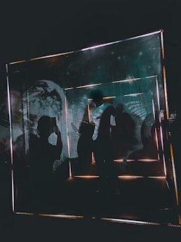 A person is silhouetted within a geometric, illuminated structure that appears to have mirrored surfaces. The scene is dimly lit with ambient and reflective light creating distorted reflections and shadows. Artwork on the walls resembles cosmic or space imagery.