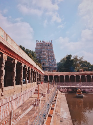 A grand temple with intricate architecture featuring a tall gopuram adorned with colorful sculptures. The temple courtyard includes a partially open area with columns and a water tank. Surrounding greenery adds a natural backdrop.