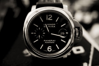 A close-up of a luxury wristwatch with a sleek black dial, featuring bold white numerals and markers. The watch face has a small seconds subdial and a date window displaying the number 26. The casing is shiny and metallic, with a crown guard visible on the side.