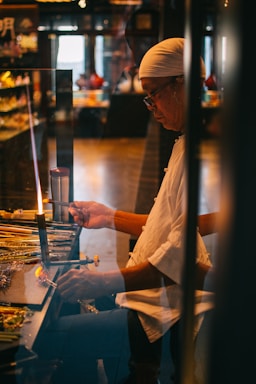 A person wearing a white headscarf and glasses is working on a craft project at a workstation. The setting appears to be a dimly lit workshop or studio, with various tools and materials laid out on the table. The individual is focused on manipulating a small tool, possibly related to glasswork or jewelry making.