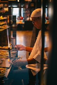 A person wearing a white headscarf and glasses is working on a craft project at a workstation. The setting appears to be a dimly lit workshop or studio, with various tools and materials laid out on the table. The individual is focused on manipulating a small tool, possibly related to glasswork or jewelry making.