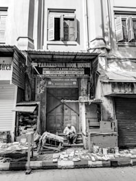 An old building facade in black and white, with open wooden shutters on the windows. Below, a bookstore named 'Tarakeswar Book House' is situated, with various books displayed on tables and the ground. A man is seated, reading a book amidst the collection. The scene appears quiet and still, capturing a moment of calm in a busy street.