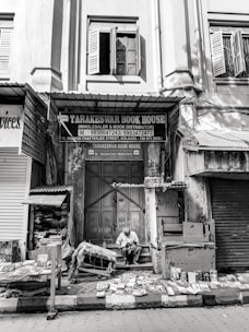 An old building facade in black and white, with open wooden shutters on the windows. Below, a bookstore named 'Tarakeswar Book House' is situated, with various books displayed on tables and the ground. A man is seated, reading a book amidst the collection. The scene appears quiet and still, capturing a moment of calm in a busy street.